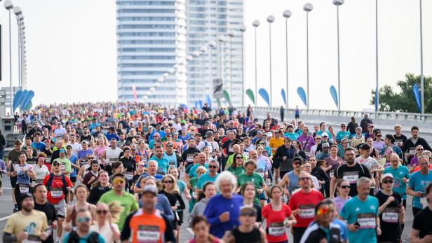 Viele Menschen laufen bei einem großen Stadtmarathon auf einer breiten Straße mit Hochhäusern im Hintergrund.