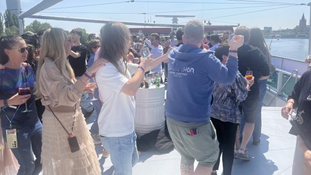 Menschen tanzen und feiern bei Sonnenschein auf einem Boot, einige halten Getränke in der Hand, im Hintergrund ist ein Fluss zu sehen.