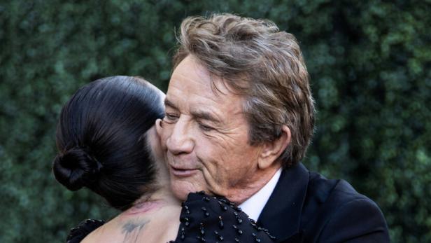 Zwei elegant gekleidete Personen - Martin Short und Selena Gomez - umarmen sich herzlich vor grünem, unscharfem Hintergrund.