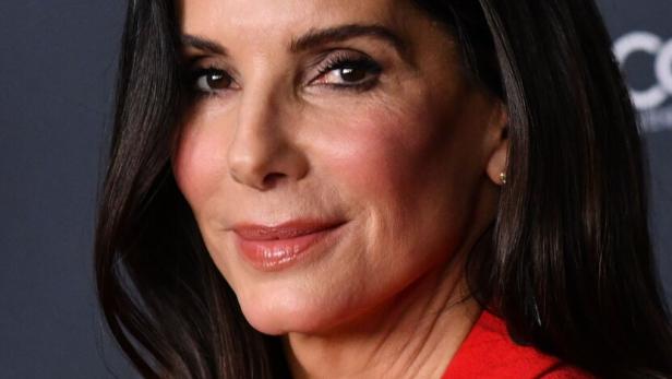 Sandra Bullock mit dunklen, gewellten Haaren und rotem Blazer vor dunklem Hintergrund.