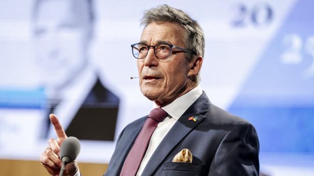 Ein Mann mit Brille und Anzug gestikuliert vor einem Mikrofon auf einer Bühne.
