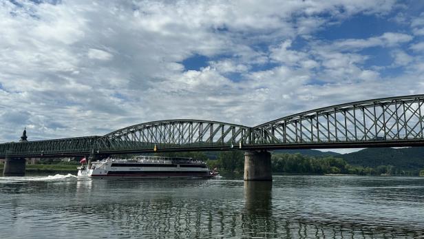 Ein Ausflugsschiff fährt unter einer großen Stahlbrücke über einen ruhigen Fluss, im Hintergrund bewaldete Hügel und Wolken.