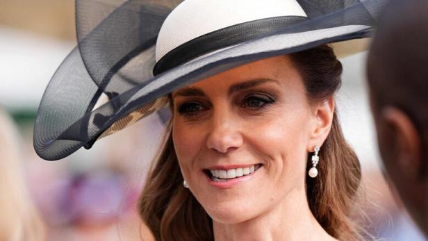 Kate trägt einen eleganten Hut mit schwarzer Schleife und ein weißes Kleid mit schwarzen Knöpfen.