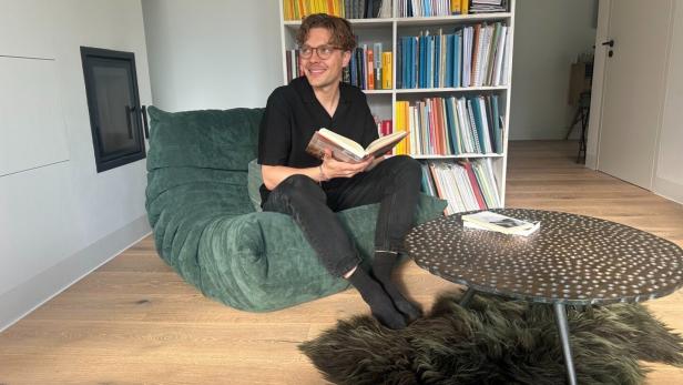 Ein Mann sitzt lächelnd mit einem Buch auf einem grünen Sessel vor einem Bücherregal und einem runden Tisch.