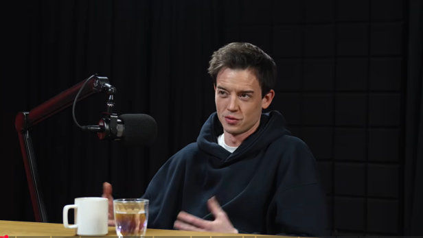 Mann spricht in einem Tonstudio vor einem Mikrofon, auf dem Tisch stehen eine Tasse und ein Glas Wasser.