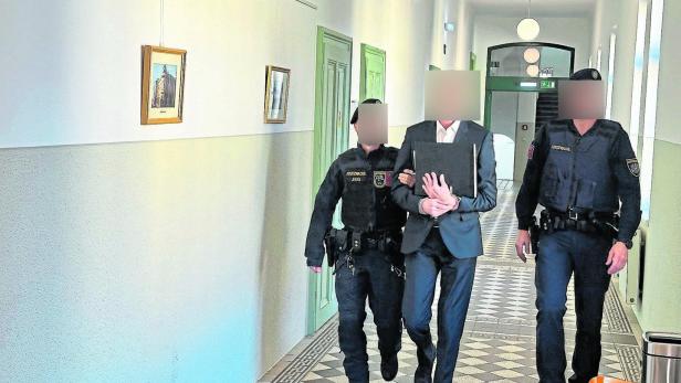 Zwei Justizbeamte führen eine Person in Handschellen durch einen hellen Flur mit grünen Türen und Bildern an der Wand.