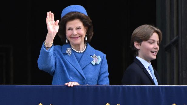 Königin Silvia winkt, begleitet von einem jungen Mann, von einem Balkon.