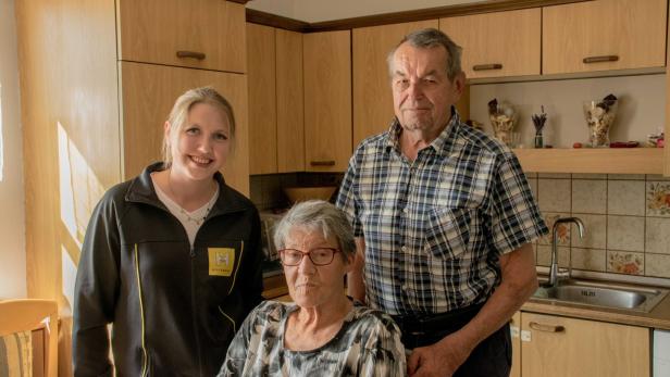 Eine junge Frau, ein älterer Mann und eine ältere Frau sitzen und stehen gemeinsam in einer Küche.
