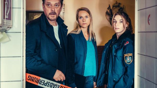 Drei Personen, darunter eine Polizistin, stehen hinter einer Absperrung mit „Polizeiabsperrung“-Band in einem Flur.
