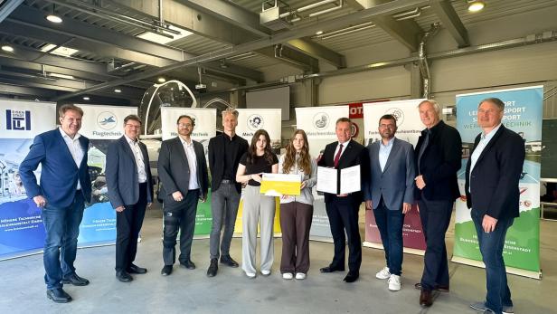 Eine Gruppe von Menschen steht in einer Halle vor Informationsplakaten, zwei junge Frauen halten eine Urkunde und einen Scheck.