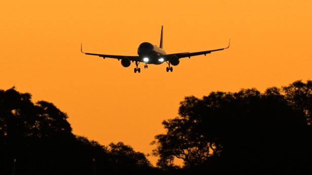 Ein Flugzeug mit eingeschalteten Scheinwerfern landet vor orangefarbenem Himmel über dunklen Bäumen.