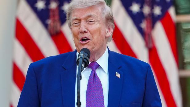Donald Trump spricht vor amerikanischen Flaggen in einem blauen Anzug und einer gemusterten Krawatte.
