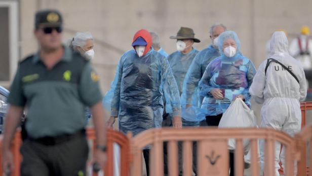 Eine Gruppe von Personen mit Masken und blauen Überwürfen wird von einem Wachmann und einem UME-Mitarbeiter begleitet.