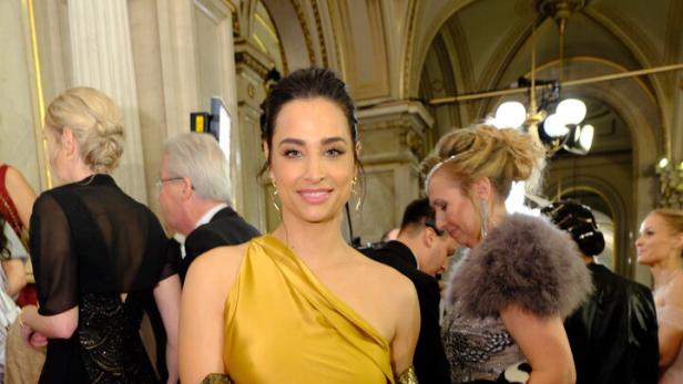 Amira Aly in einem goldenen, schulterfreien Kleid posiert auf einem roten Teppich.