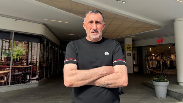 Ein Mann mit verschränkten Armen steht vor einem Einkaufszentrum mit H&M-Eingang im Hintergrund.