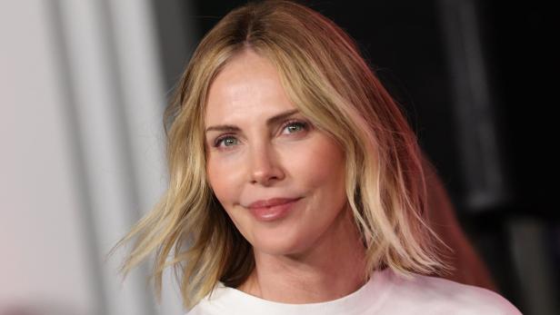 Charlize Theron mit blonden, gewellten Haaren und einem weißen T-Shirt lächelt.