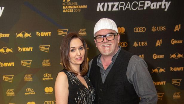 Eine Frau im eleganten schwarzen Kleid und ein Mann mit Mütze und Brille posieren gemeinsam auf einem gelben Teppich.