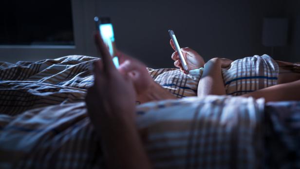 Zwei Personen liegen nebeneinander im Bett und benutzen jeweils ein Smartphone im Dunkeln.