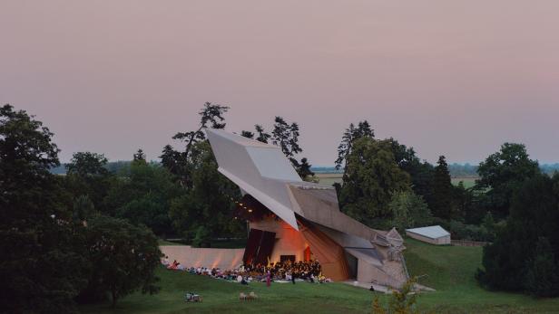 Eine moderne Freilichtbühne mit ungewöhnlicher Dachkonstruktion steht inmitten einer grünen Parklandschaft.