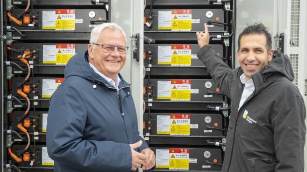 Zwei Männer stehen vor mehreren großen Batteriespeichern mit Warnhinweisen und lächeln, einer zeigt auf eine Batterie.