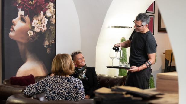 Ein Mann schenkt zwei Frauen in gemütlicher Vinothek Wein ein, im Hintergrund ein großes Porträt.