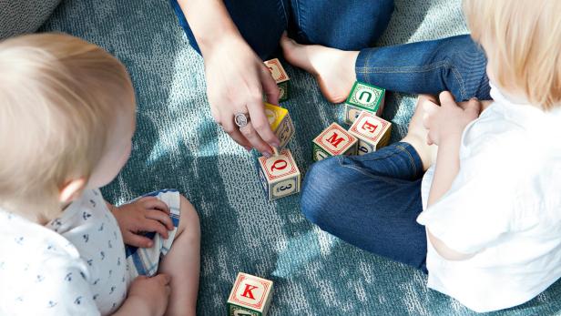 Zwei kleine Kinder und eine erwachsene Person spielen gemeinsam mit bunten Holzbausteinen auf dem Teppich.