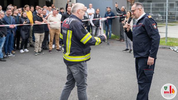 Zwei Männer in Feuerwehrkleidung sprechen vor einer größeren Menschenmenge im Freien, einer hält ein Mikrofon.