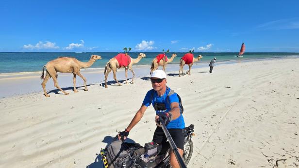 Ein Mann macht ein Selfie auf einem Fahrrad am Strand, während mehrere Kamele mit roten Decken am Meer entlanggeführt werden.