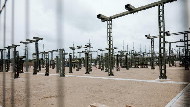 Viele grüne Metallgerüste stehen auf einem sandigen Gelände, im Hintergrund sind Strommasten und Bauzäune zu sehen.