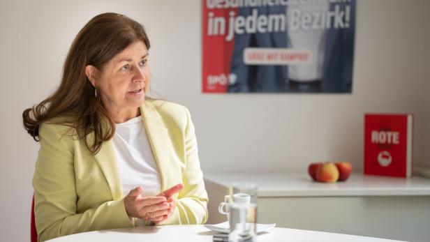 Eine Frau im hellgelben Blazer sitzt an einem Tisch, im Hintergrund ein SPÖ-Plakat.