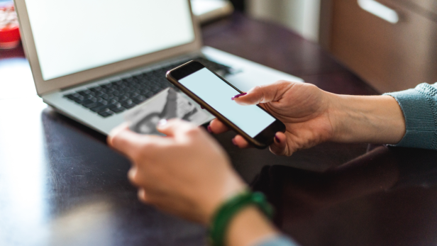 Eine Person nutzt Smartphone und Kreditkarte an einem Laptop für einen Online-Kauf.