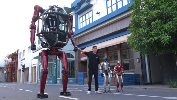 Unitree-CEO Xingxing Wang steht neben dem neuen, rot lackierten Mecha GD01 und zwei kleineren humanoiden Robotern auf einer Straße.