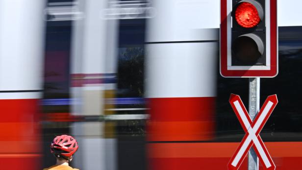 Ein Kind mit Fahrrad wartet an einem Bahnübergang mit rotem Signal, während ein Zug vorbeifährt.