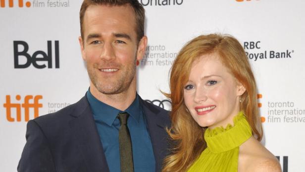 James Van Der Beek im Anzug und seine Ehefrau Kimberly Van Der Beek im grünen Kleid stehen lächelnd nebeneinander vor einer Sponsorenwand. Aufgenommen wurde das Foto im Jahr 2013.