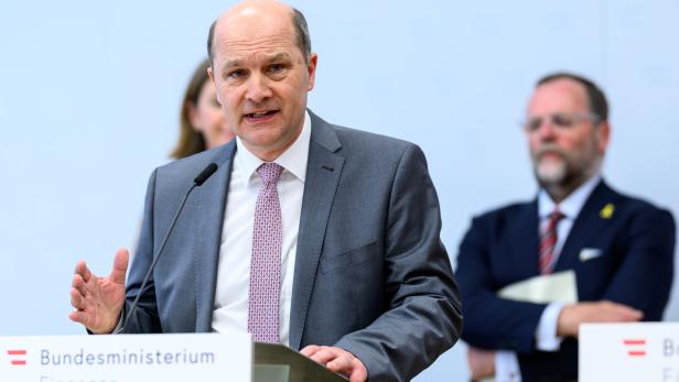 Ein Mann im Anzug spricht an einem Rednerpult des Bundesministeriums für Finanzen, im Hintergrund stehen zwei Personen.