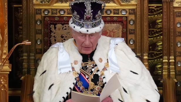 König Charles III. mit Krone und Hermelinmantel liest von einem Blatt Papier auf einem goldenen Thron.