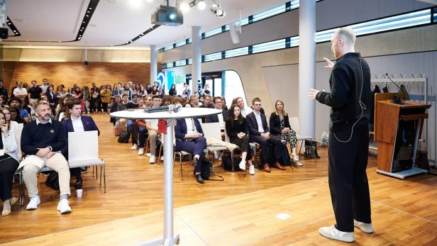 Ein Mann hält einen Vortrag auf einer Bühne vor einem aufmerksam zuhörenden Publikum in einem modernen Saal.