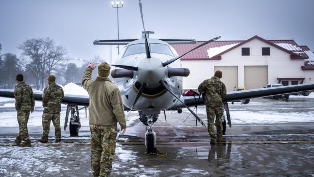 Vier Soldaten in Tarnuniformen inspizieren ein Propellerflugzeug auf einem verschneiten Flugplatz.