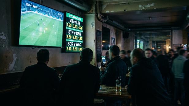 Dunkle Bar mit mehreren Personen, die auf eine große Wandbildschirm mit Fußballspiel und daneben angezeigten Wettquoten schauen