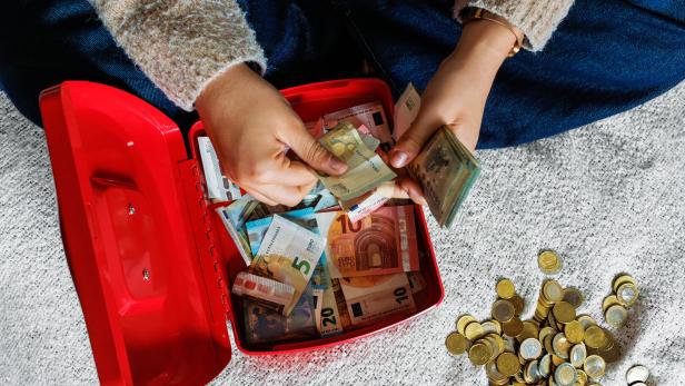 Eine Person zählt Geldscheine aus einer roten Kasse, daneben liegt ein großer Haufen Euromünzen auf dem Teppich.