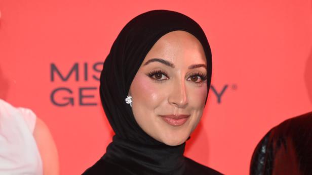 Eine Frau mit schwarzem Hijab lächelt vor einem roten Hintergrund.