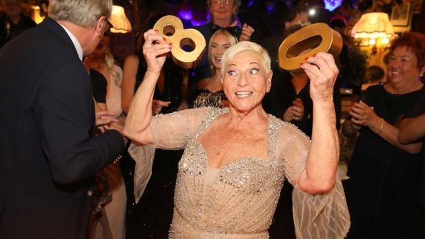 Eine strahlende Frau hält goldene Zahlen über einem Geburtstagskuchen mit Kerzen.
