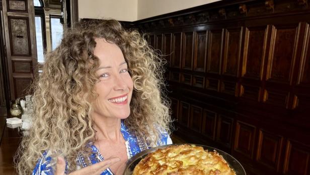 Eine Frau mit lockigem Haar präsentiert lächelnd einen runden Kuchen auf einem silbernen Tablett.