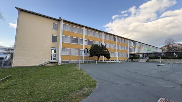 Ein langes, gelbliches Schulgebäude mit vielen Fenstern steht neben einem leeren Schulhof mit etwas Grünfläche und einem Baum.
