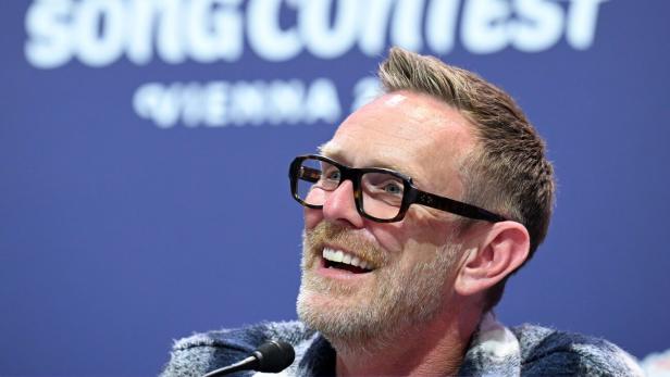 Ein Mann mit Brille lächelt vor einem Hintergrund mit dem Schriftzug „Eurovision Song Contest Vienna“.