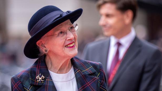 Königin Margrethe II. lächelt in einem karierten Blazer und Hut, während ein Mann im Anzug im Hintergrund steht.