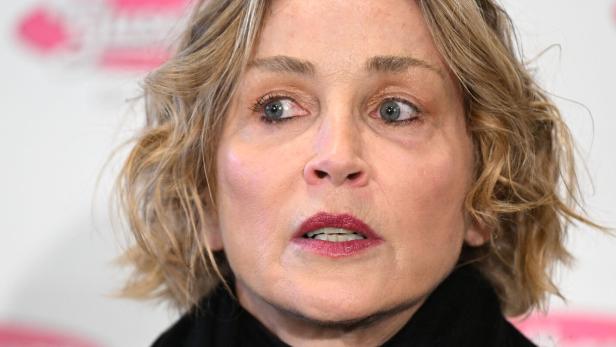 Sharon Stone mit blonden, gewellten Haaren und dunklem Rollkragenpullover blickt aufmerksam zur Seite.