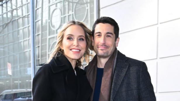 Jason Biggs und Jenny Mollen stehen eng umarmt auf einem Gehweg vor einem modernen Gebäude, beide tragen Wintermäntel.