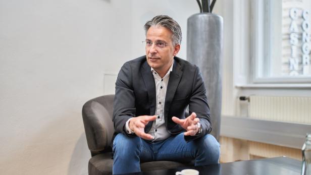 Ein Mann mit Brille, Sakko und Jeans sitzt gestikulierend in einem Sessel.