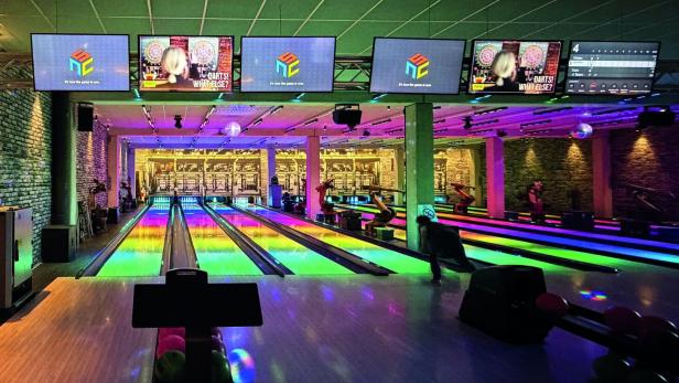 Mehrere Bowlingbahnen mit bunten Lichtern, Menschen beim Bowling und Bildschirmen über den Bahnen.